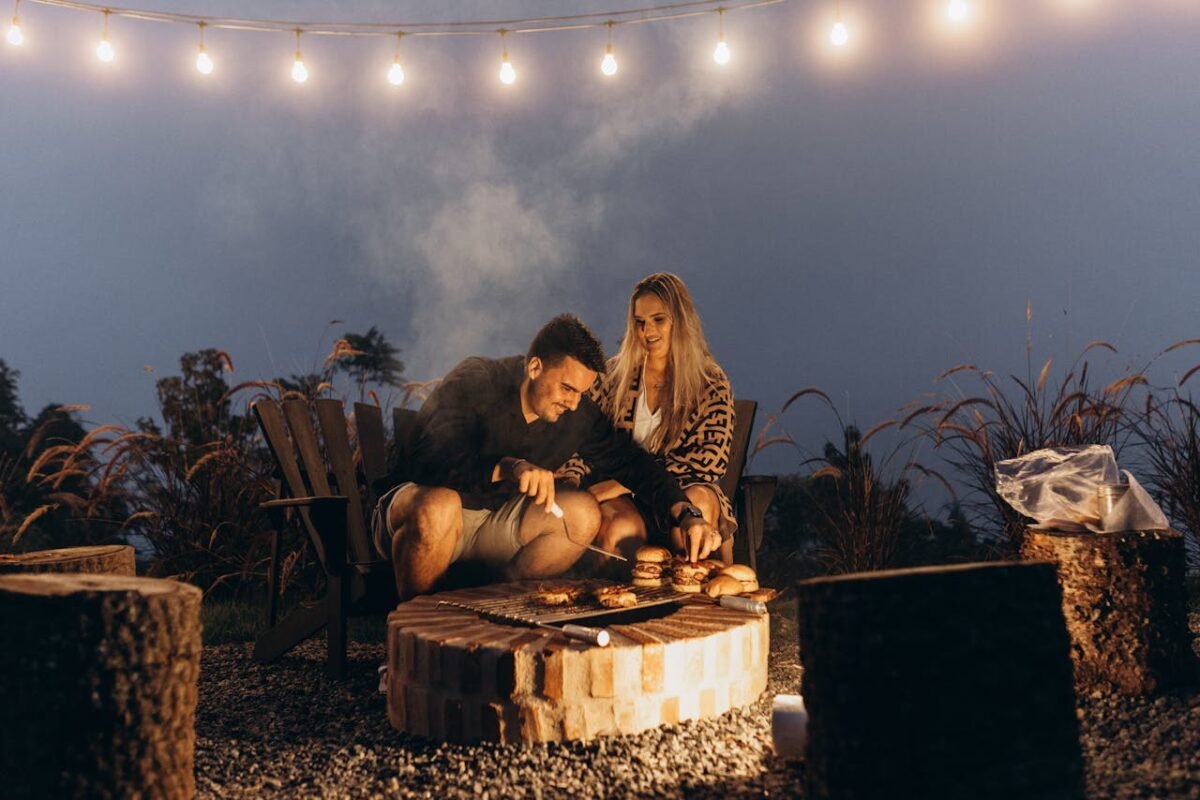 Couple enjoying a cozy barbecue by a fire with glowing string lights, creating a warm, romantic evening.
