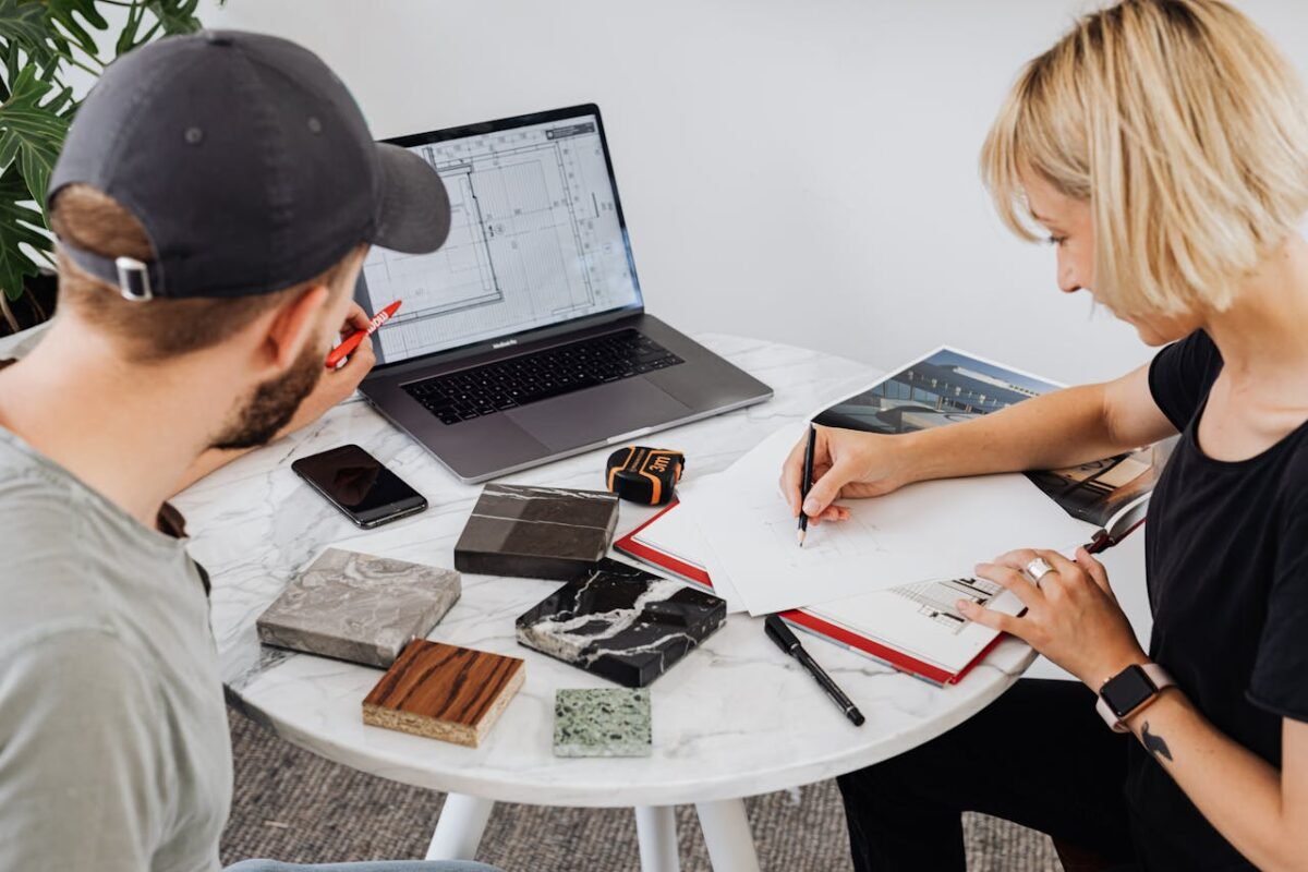 Two designers collaborating on a project, utilizing a laptop and drafting materials on a round table.