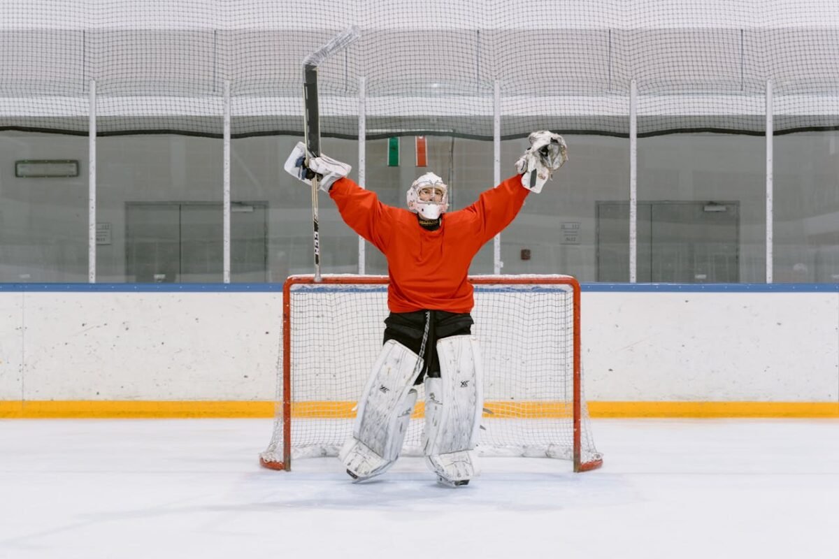 Ice hockey goalie in red jersey celebrating with raised arms in the rink's goal area.