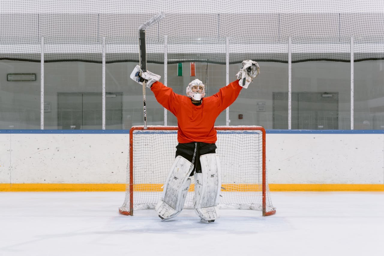 Ice hockey goalie in red jersey celebrating with raised arms in the rink's goal area.