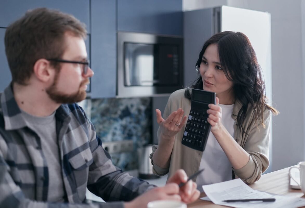 A couple sitting in a kitchen discussing financial matters using a calculator and documents.